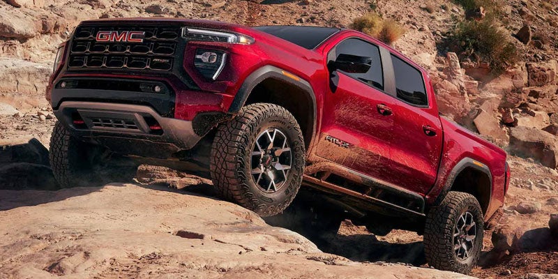 Red GMC pickup truck with large off-road tires climbing over rocky terrain.