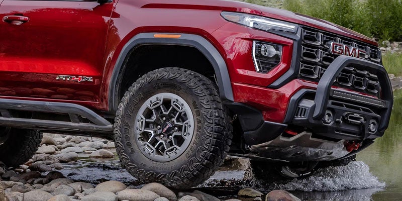 GMC truck with large off-road tire and heavy-duty bumper on rocky terrain, splashing water.