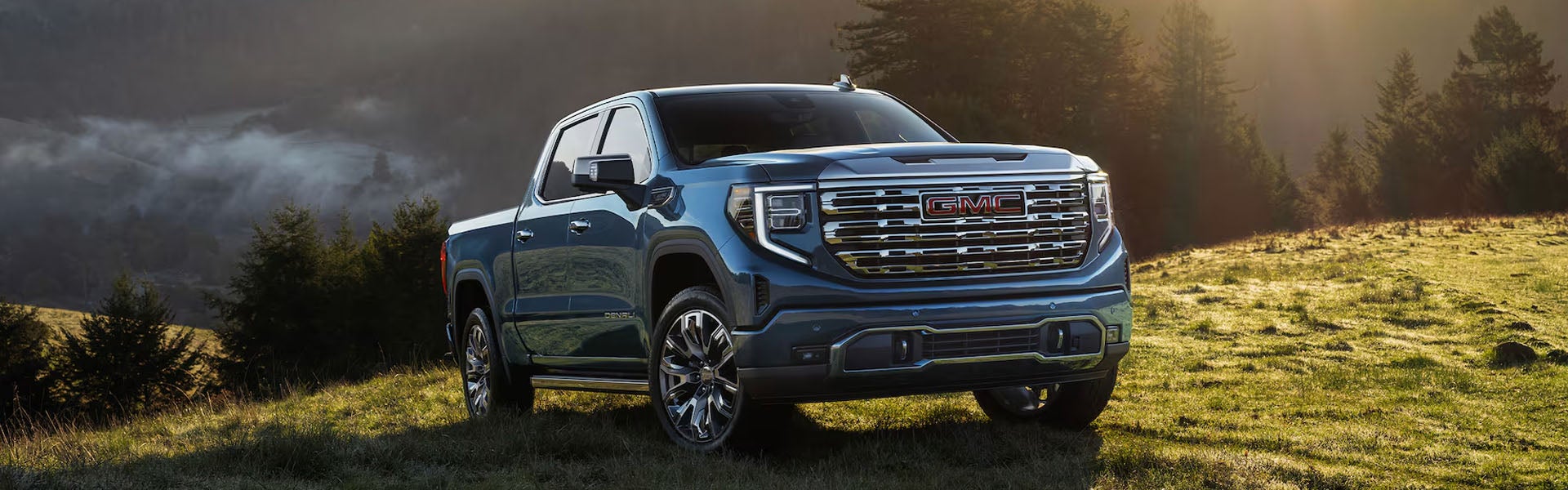GMC pickup truck parked on a grassy hill with sunlit trees and misty mountains in the background.