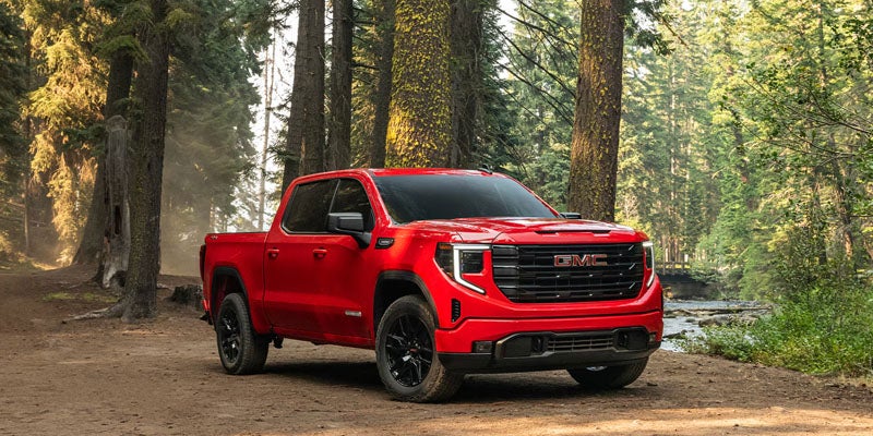 GMC pickup truck parked on a dirt path in a forest with tall trees and a stream nearby.