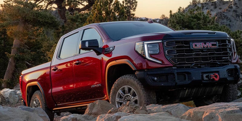 Red GMC AT4X pickup truck driving over rocky terrain with trees and mountains in the background.