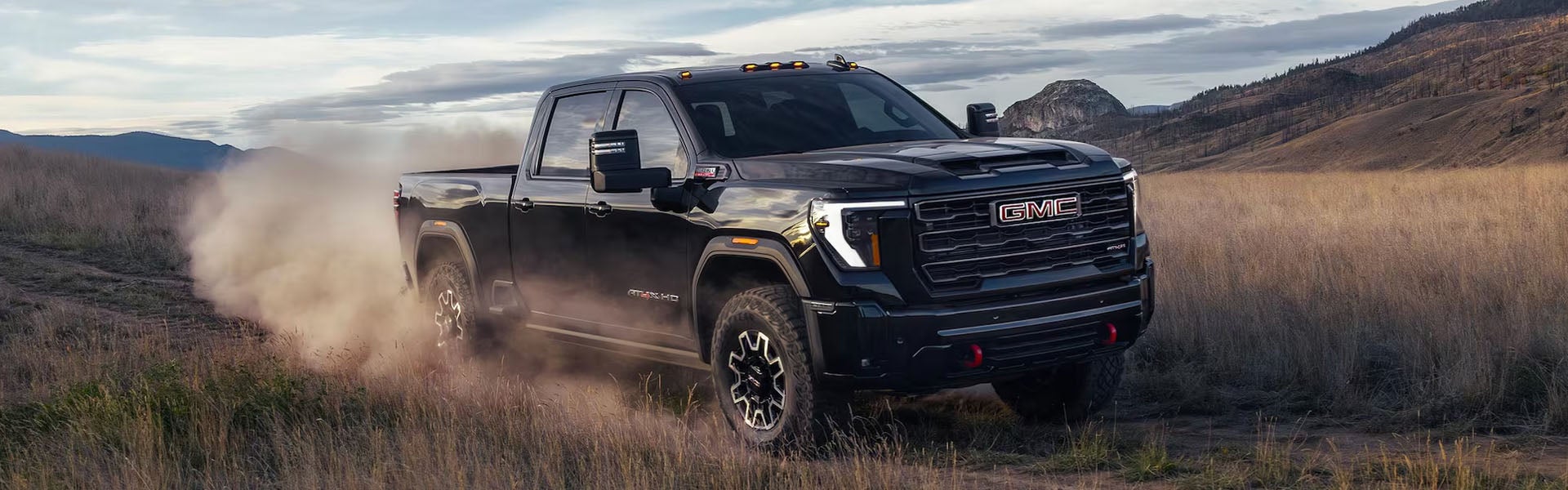 GMC truck driving on a dirt path, kicking up dust in a dry, grassy landscape.