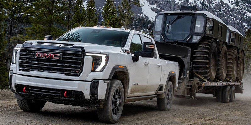 White GMC pickup truck towing a large, multi-wheeled amphibious vehicle on a flatbed trailer on a dirt road.