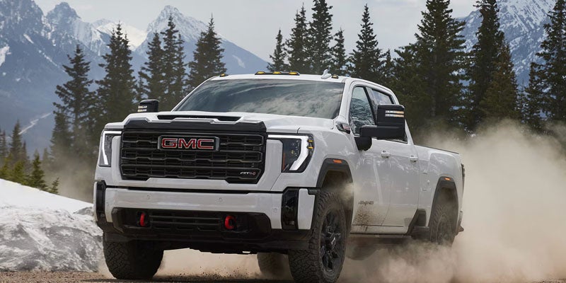 GMC pickup truck driving on a dirt road, kicking up dust, with snowy mountains and pine trees in the background.
