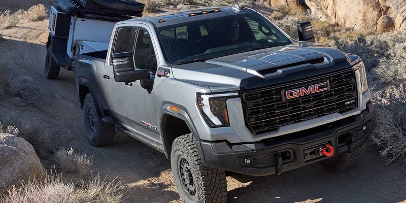 Silver GMC pickup truck with rugged tires towing an off-road trailer on a dirt path in a rocky, arid landscape.