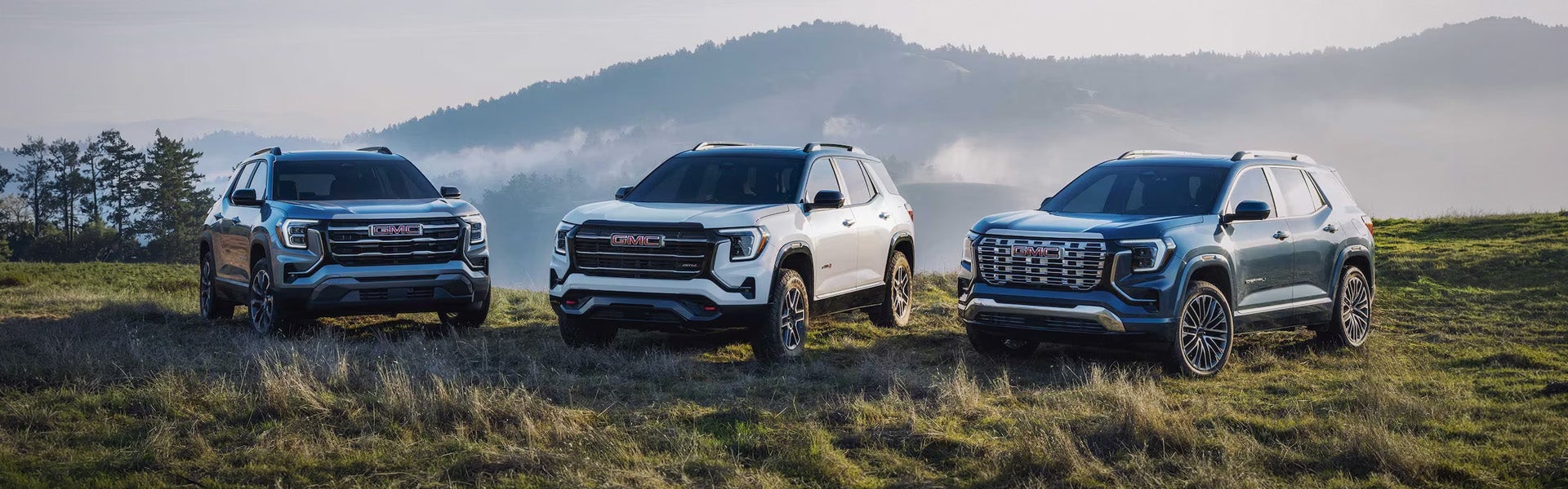 GMC SUVs (dark blue, white, and light blue) parked on a grassy hill with misty mountains in the background.