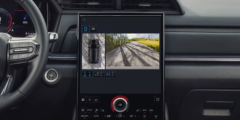 Large touchscreen in a car dashboard shows split-screen camera views: a dashcam and a dirt road with trees.