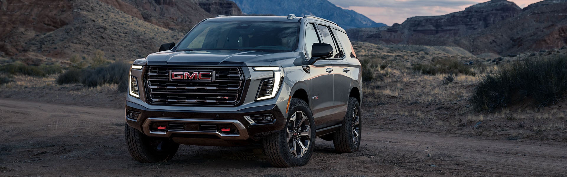 Grey GMC Yukon AT4 SUV on a dirt road in a desert landscape with mountains under a cloudy sky.