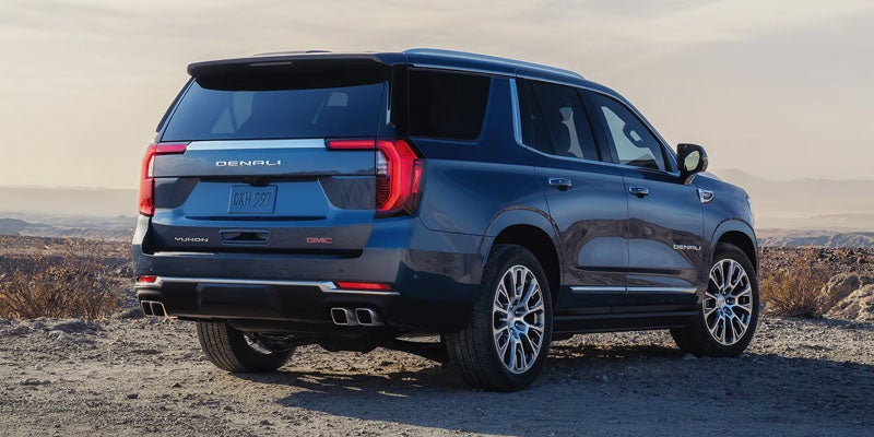 Dark blue GMC Yukon Denali SUV, rear view, parked on a dirt road with a hazy desert landscape behind it.