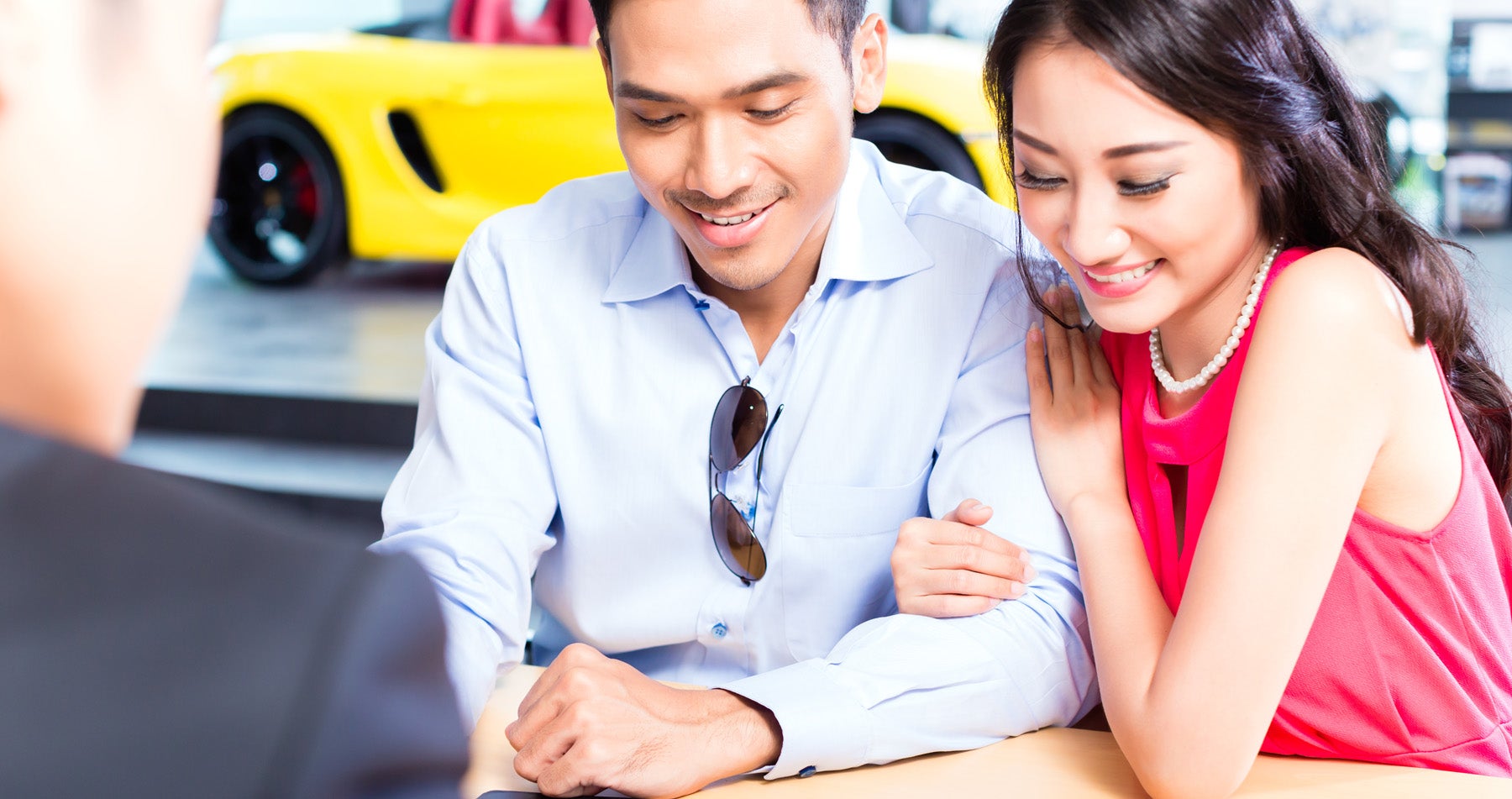 A smiling couple looks at a yellow sports car while standing in a car dealership.