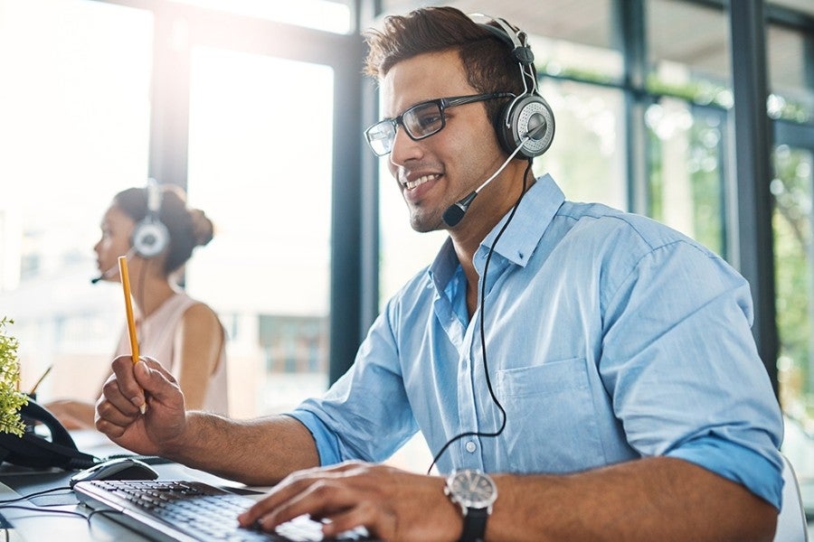 A man with a headset smiles while typing on a computer at his desk.