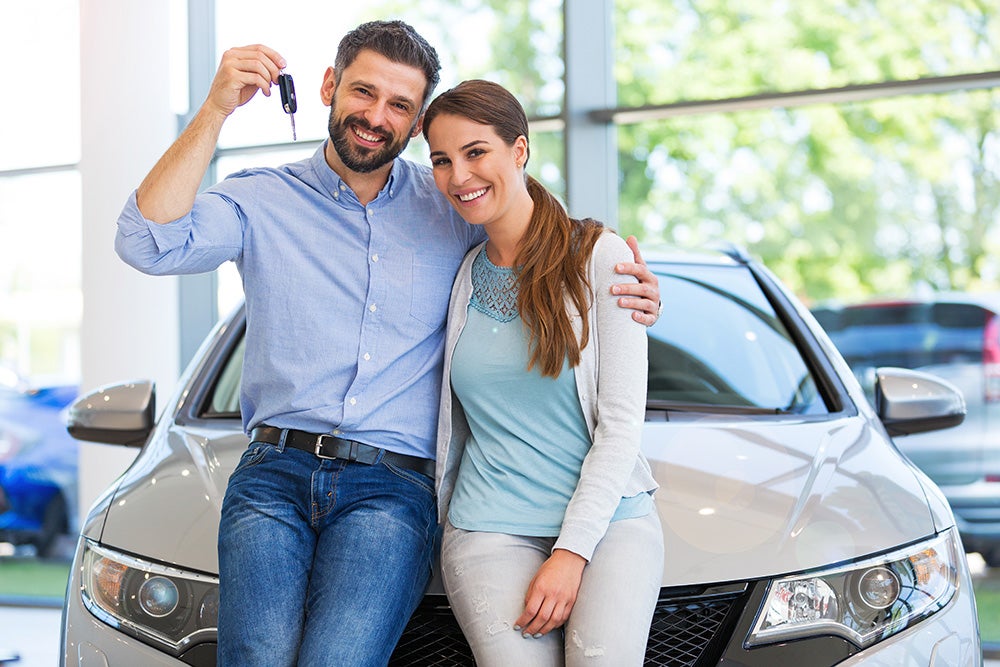 A smiling couple holds car keys and poses in front of a new gray vehicle.