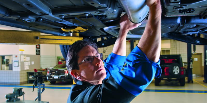 A mechanic wearing glasses holds a silver car part underneath a vehicle's undercarriage.