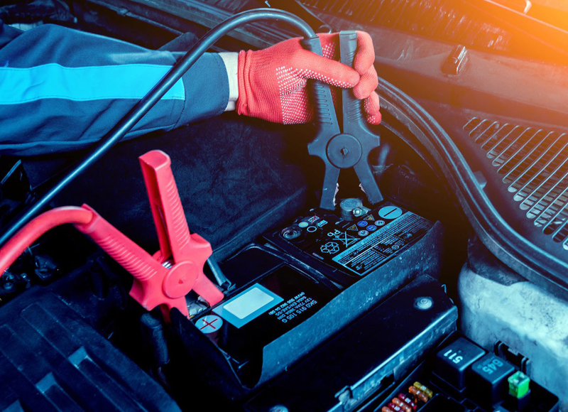 A gloved hand connects jumper cables to a car battery to jump-start a vehicle.