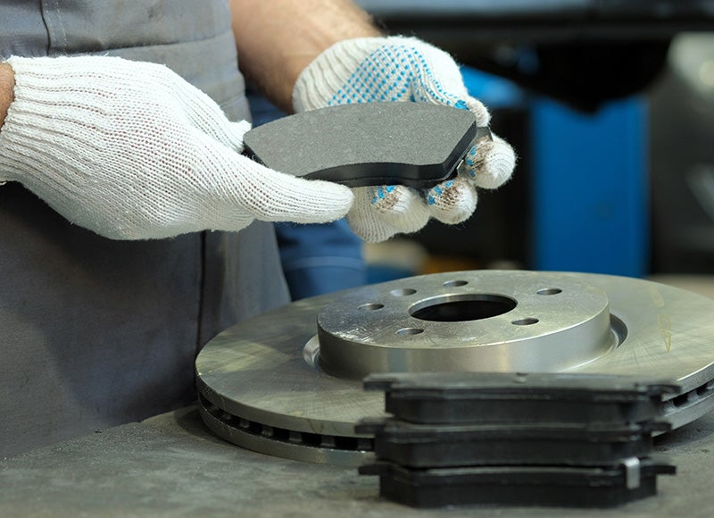 A mechanic wearing gloves holds a brake pad above a brake rotor and other pads on a workbench.