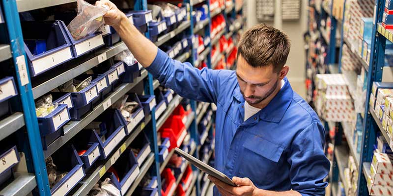 A man in a blue uniform reaches for parts in a well-stocked warehouse, looking at a tablet.