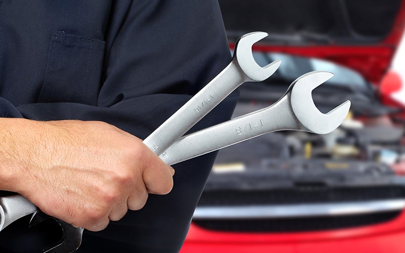 A mechanic holds a pair of wrenches in front of an open car engine.